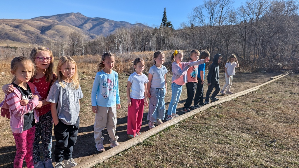 A group of elementary students lined up and balancing on a long wooden beam laid on the ground, with mountains in the background.