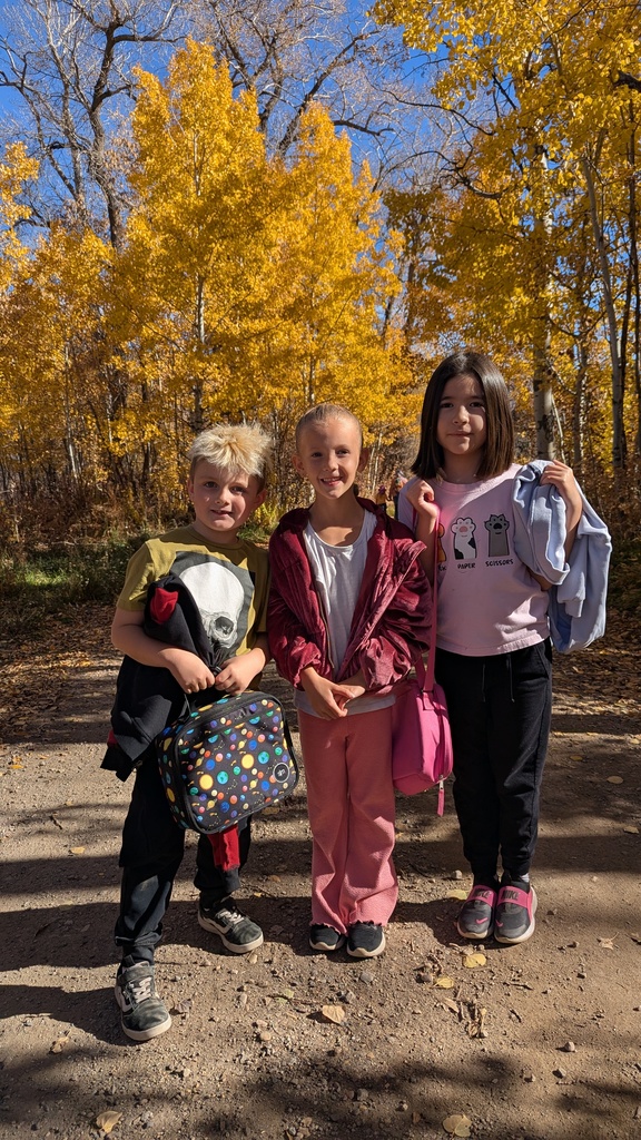 Three elementary students smiling as they stand together on a dirt path in front of a brilliant background of yellow autumn trees.