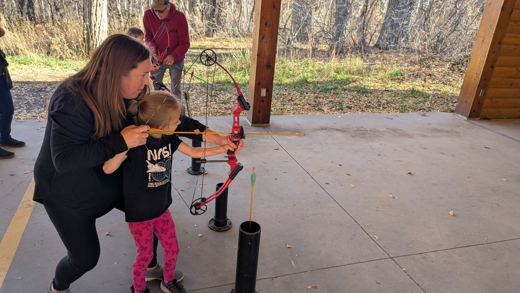 An adult chaperone assisting a young girl with a ponytail in aiming a bow and arrow at an outdoor covered range.