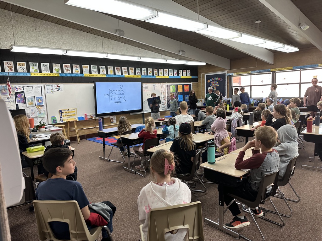 Students in a classroom setting watching a presentation. A group of older students or guest speakers are standing near a large screen displaying a diagram, while elementary-aged students sit at desks watching and listening. The room is bright and decorated with educational posters.