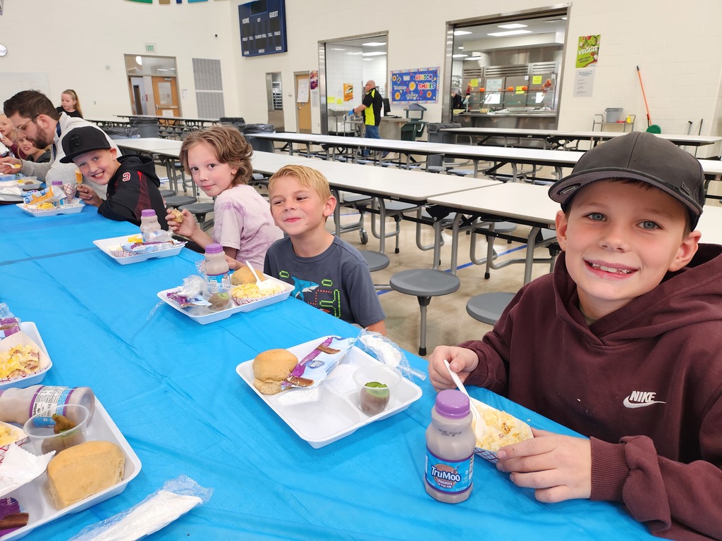 Four young boys are sitting side-by-side at a blue-covered table, smiling while eating lunch. The boy on the far right wears a black baseball cap and a maroon hoodie. In the background, a cafeteria serving line with a sign reading "VEGGIE" is visible, along with a blue scoreboard on the wall.