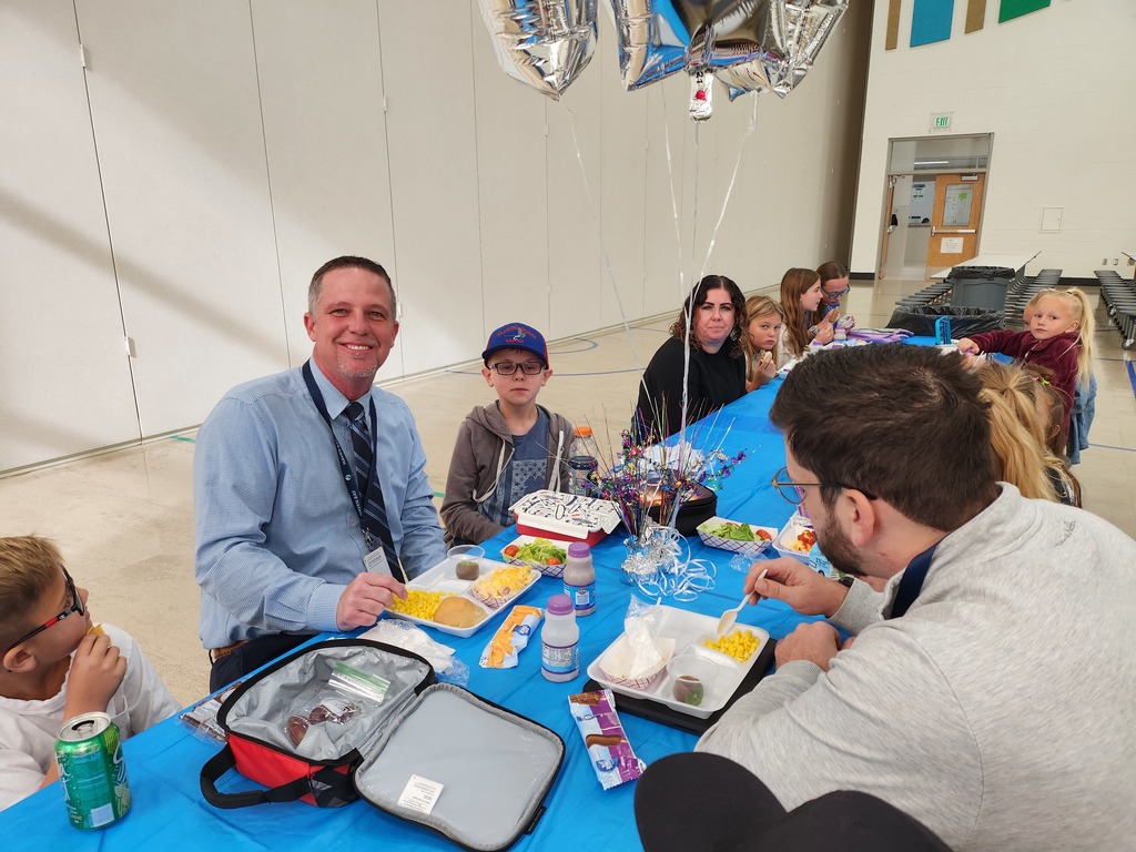 A close-up view of a group of children and two adult men seated around a blue-covered table. Justin Skeen, in a light blue patterned button-down shirt, smiles directly at the camera. Trays of food and a small, colorful, decorated item with birthday candles are on the table.