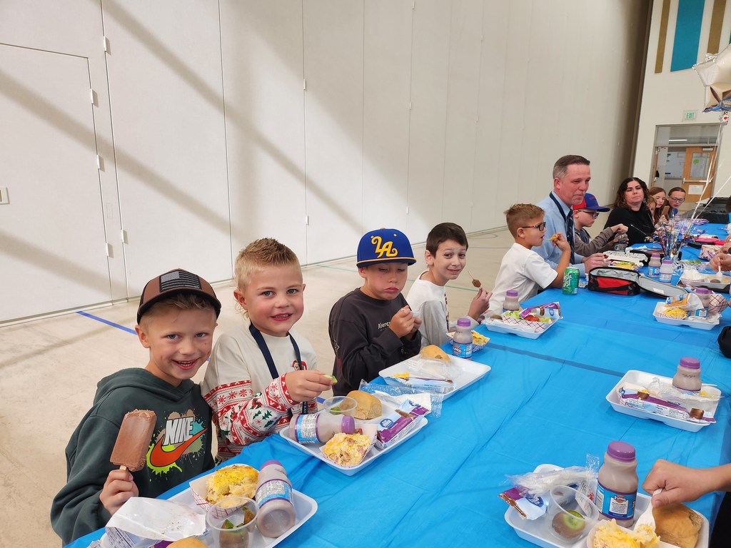 Four young boys sit at a blue-covered table, smiling and holding their lunches. The boy on the far left holds a chocolate-covered ice cream bar. Several adults, including Mr. Skeen and Mrs. Pulsipher, and other children are visible further down the long table.
