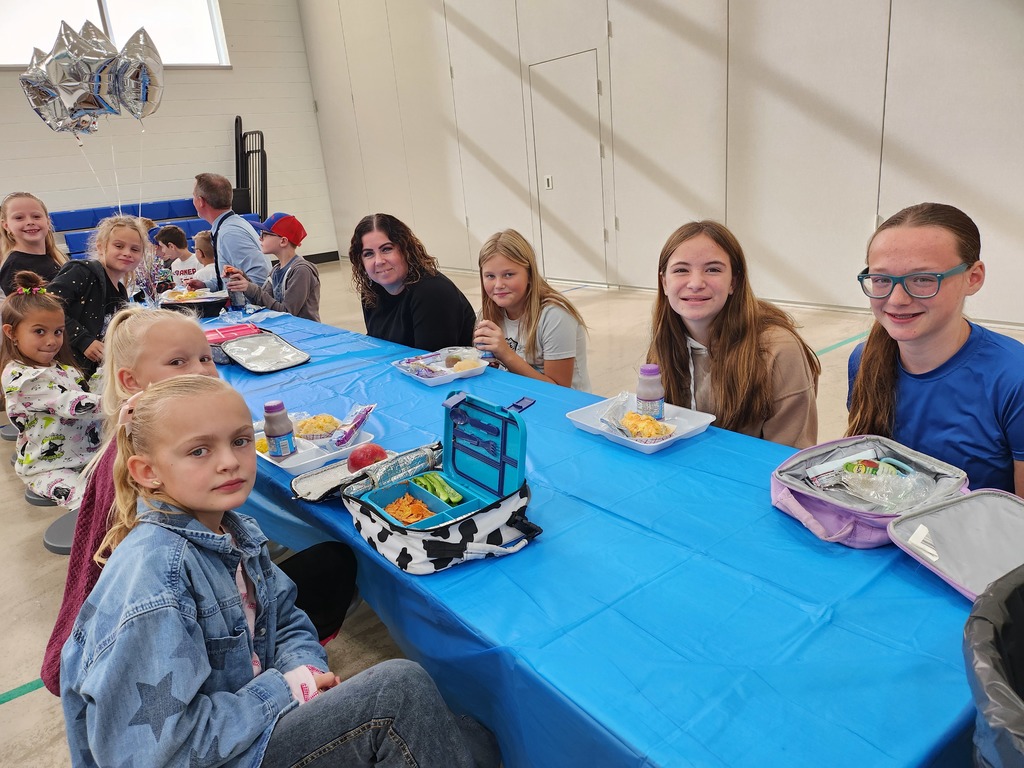 A line of approximately nine girls and a woman sitting along one side of a long, blue-covered table in a cafeteria setting. Most of the girls are smiling at the camera, and lunch trays and bags are visible on the table.