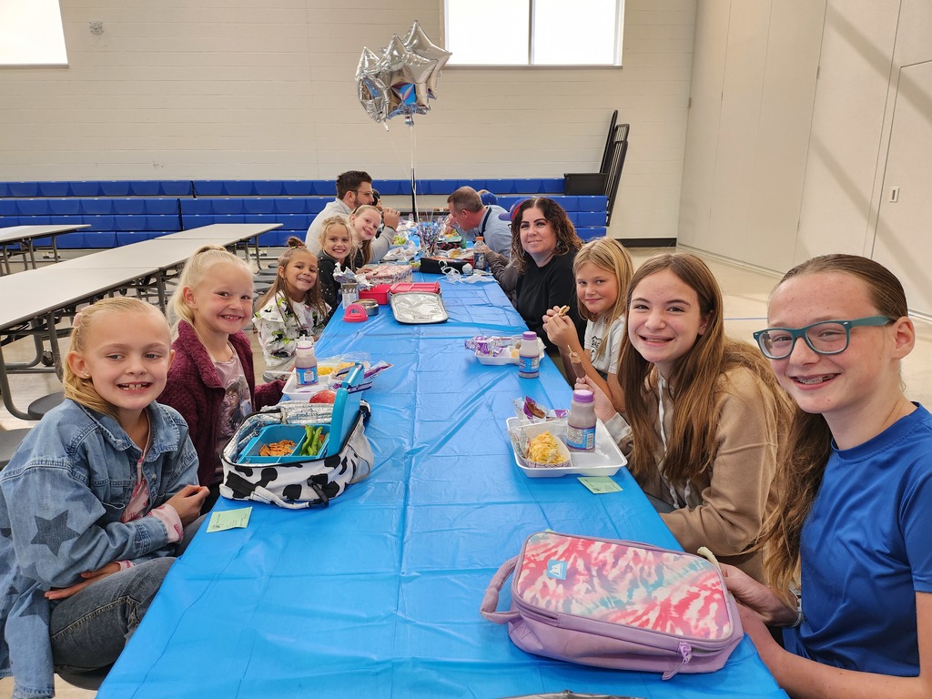 A group of approximately ten girls and a few adults sit together at a long table covered with a bright blue disposable cloth in a school cafeteria or gym. The girls in the foreground are smiling at the camera, with lunches and lunchboxes scattered across the table. Silver star-shaped balloons float above the group.