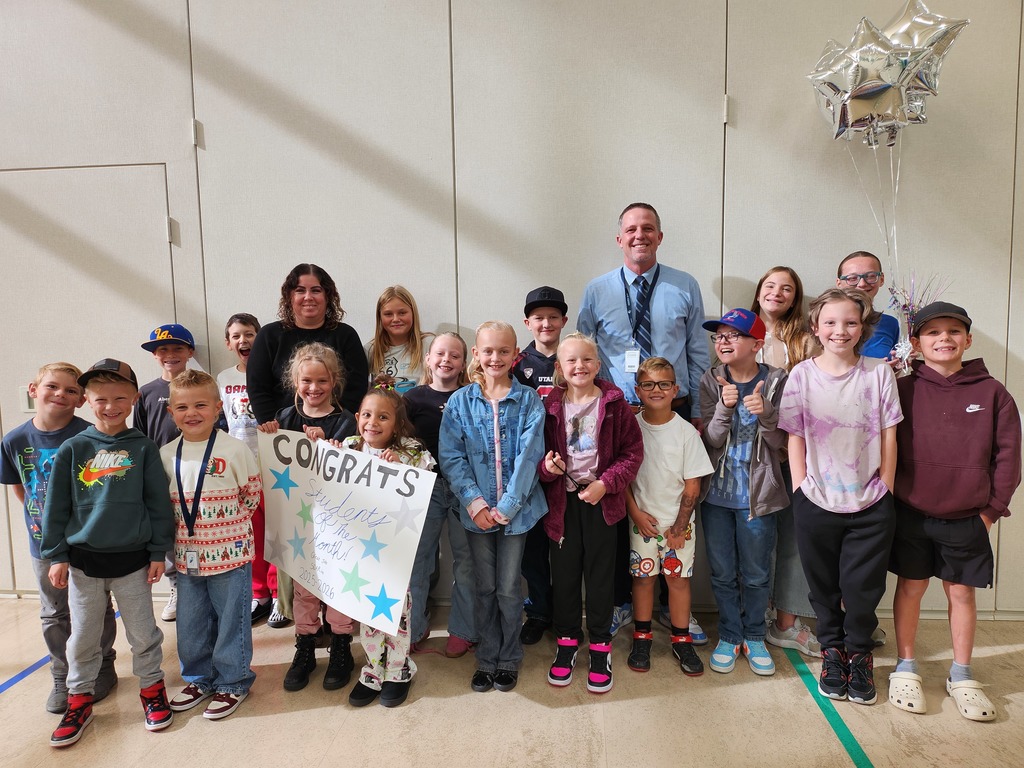 A large group photo of approximately fifteen children and two adults, Mr. Skeen and Mrs. Pulsipher, standing against a light-colored wall in a gym or cafeteria. They are posing with a sign that reads, "CONGRATS Students of the Month 2023-2024". Silver star balloons are on the right, and everyone is smiling.