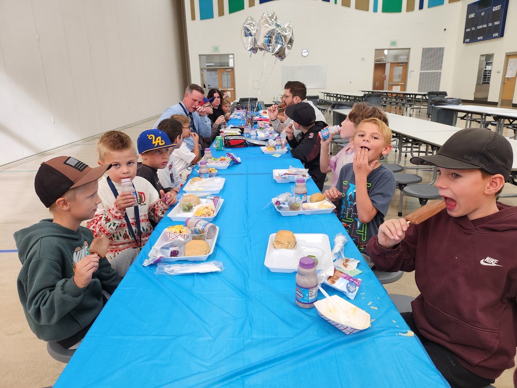 A side view of children and adults eating together at a long, blue-covered table. A young boy in the foreground on the right is taking a bite of a chocolate-covered ice cream bar. Another boy in a red and white patterned sweater is drinking from a small bottle.