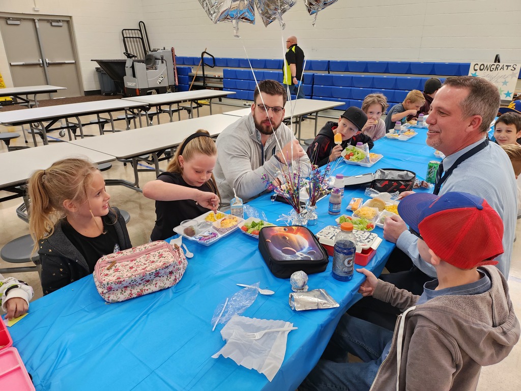 A group of children and two adult men sitting around a blue-covered table, eating lunch. Mr. Skeen in a light blue patterned shirt smiles toward the right. Two girls on the left are focused on their food. Silver star-shaped balloons are visible, and part of a banner reading "CONGRATS" is on the right.