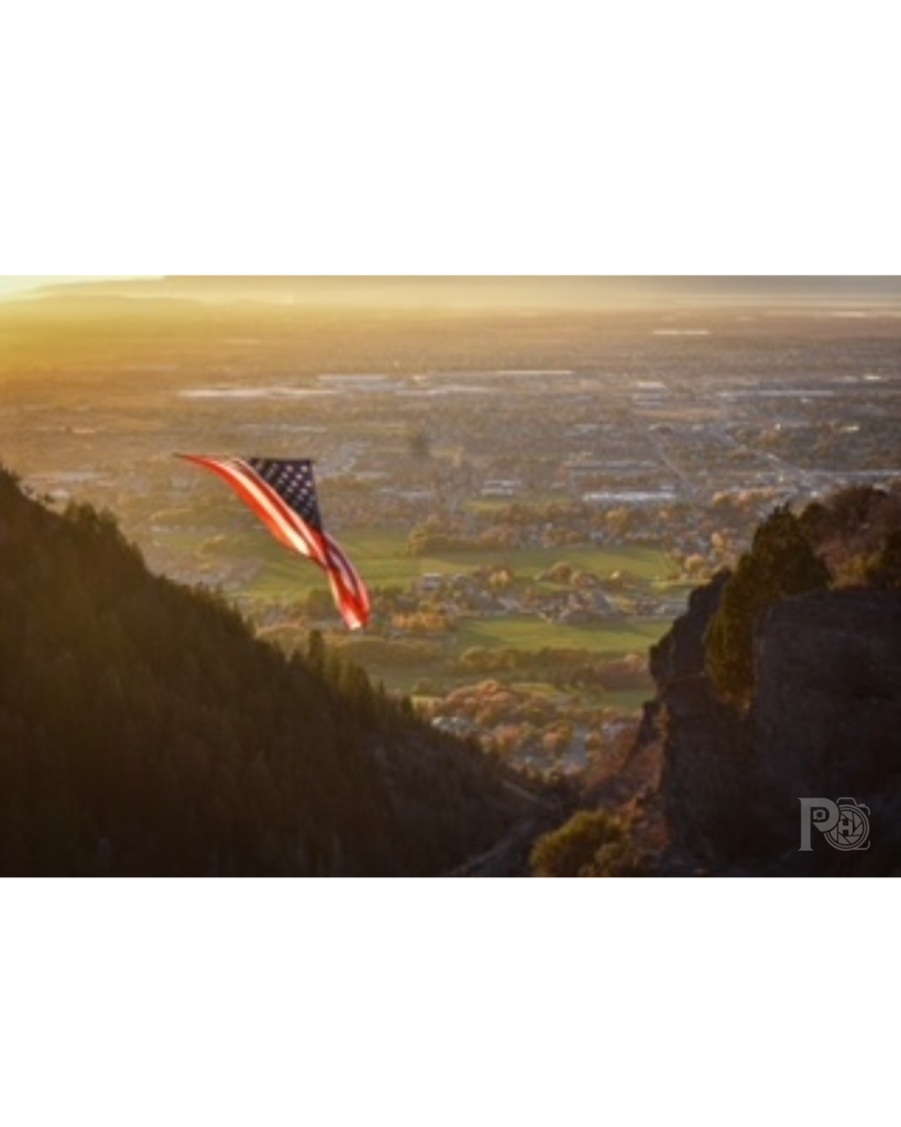 Photo of the American Flag named "The Major" hanging between the mountains of Coldwater Canyon with the Town of North Ogden City below.