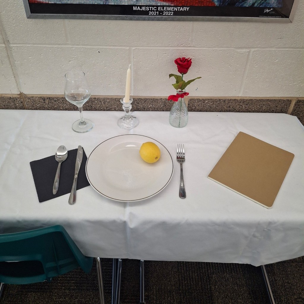 close up of white table set with plate with a lemon and salt on it. a black napkins, white candle, notebook, upside down glass, and red rose