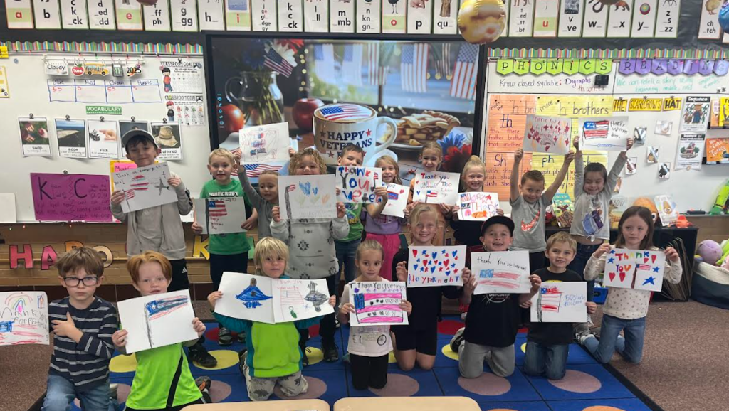 A group of approximately 15 elementary school children, smiling and kneeling or standing in a classroom. They are holding up handmade drawings and thank you notes dedicated to veterans, featuring American flags, stars, and patriotic messages. The classroom has a whiteboard with "Happy Veterans" imagery, phonics lessons, and vocabulary words visible in the background.