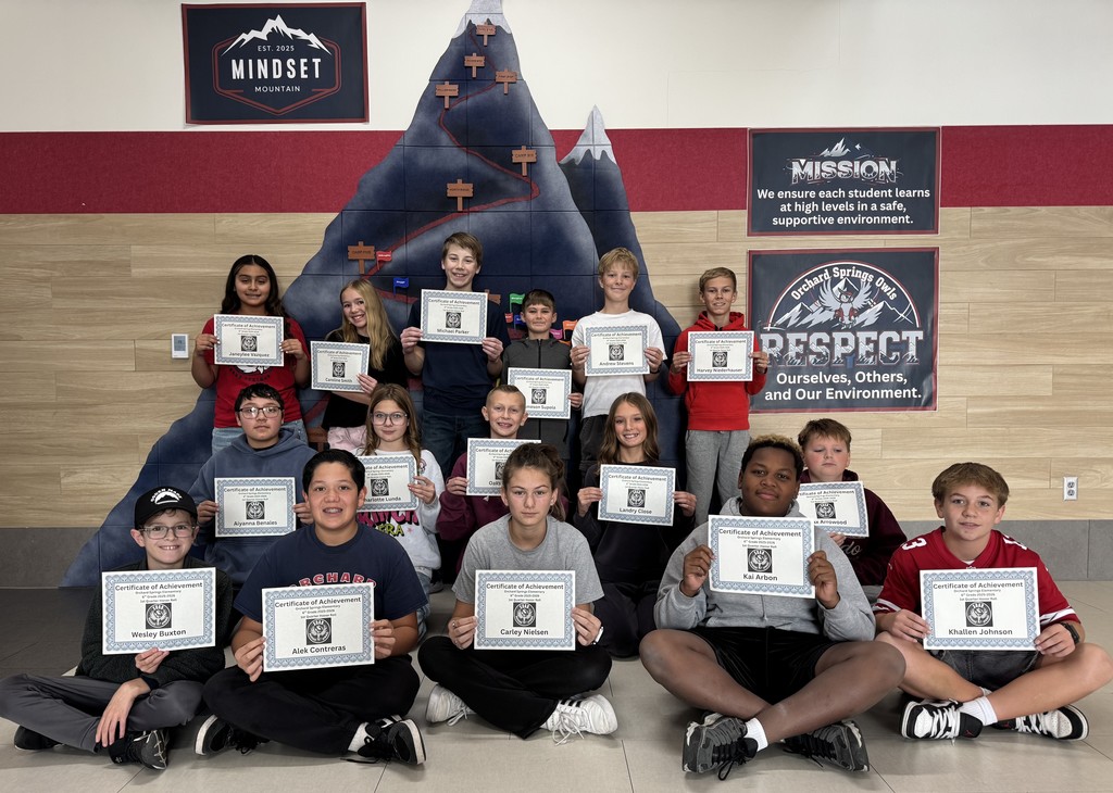 Here is a deA large group of smiling sixth-grade students posing in four tiered rows in a school hallway. Most of the students are holding up certificates of achievement. Behind them is a wall featuring a large mural of a mountain labeled 'Mindset Mountain' and a poster for 'Orchard Springs Owls' with a mission statement. The group represents the High Honor Roll recipients for the first quarter.