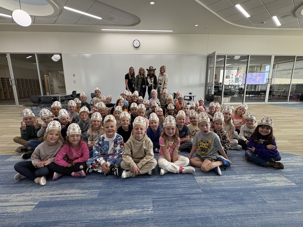 A large group of approximately 50 elementary school children are sitting close together on a blue and tan patterned carpet in a bright, modern room with white walls and a distinctive ceiling. Most of the children are wearing identical white paper hats/caps with a brown bear design. Four adult women, one of whom is wearing a cowboy hat, are standing in the back center against the white wall.