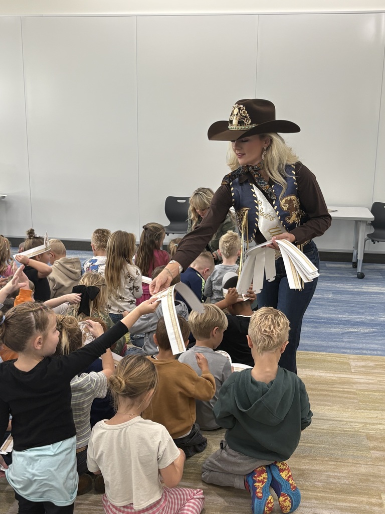 A blonde woman in a cowboy hat and rodeo attire (dark shirt, patterned vest, jeans) bends down to hand out thin, white, folded items (possibly paper strips or bookmarks) to a large group of young children sitting cross-legged on a carpeted floor. The children are reaching up to receive the items.