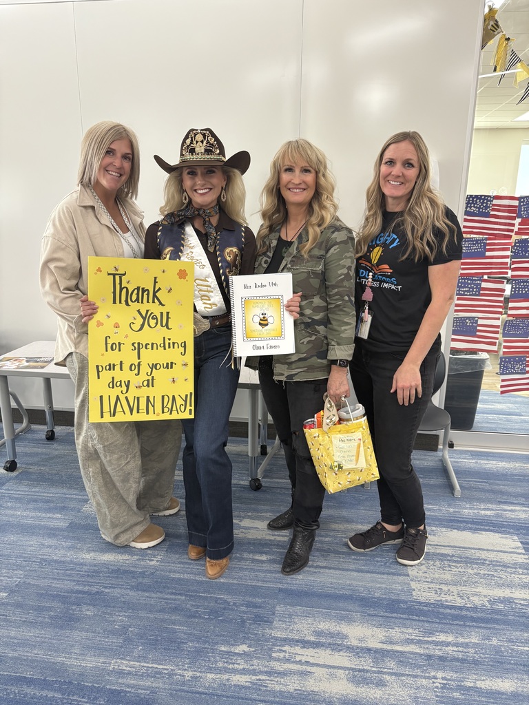 Four women are standing side-by-side in a modern room with a blue and tan carpet. The second woman from the left is wearing a cowboy hat and rodeo attire, including a sash that says "Utah" on it. The two women on the left are holding a large yellow sign that reads, "Thank You for spending part of your day at HAVEN BAY." The woman next to the cowboy is holding a card that reads, "The Little Bear. Clint Swin." The woman on the far right is wearing a black t-shirt that reads "FLIGHT NIGHT DIFFERENCE MAKERS IMPACT" and is holding a yellow gift bag. The background includes a display of small American flags.