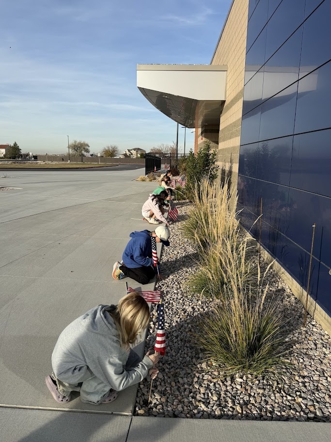 Several children are kneeling near a flowerbed next to the school building, carefully placing small American flags into the ground.