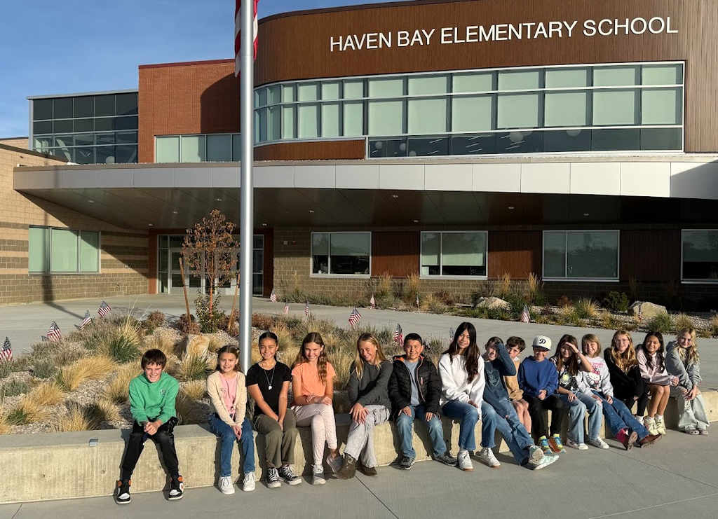 A group of school-aged children are sitting on a ledge in front of the HAVEN BAY ELEMENTARY SCHOOL building. A flagpole with an American flag stands behind them, and small American flags are planted in the landscaping.
