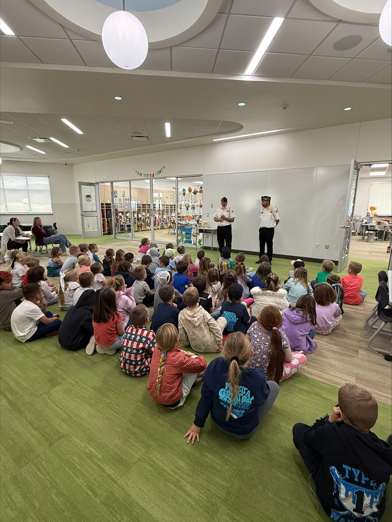 Two men in Veterans of Foreign Wars (VFW) uniforms are standing in front of a large group of young children sitting on a green carpet in a bright, modern room, apparently giving a presentation.