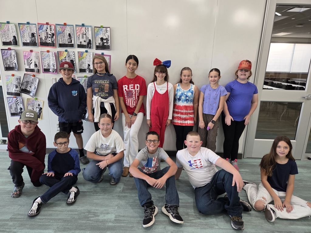 A group of approximately 13 fifth-grade students is posing indoors, many wearing red, white, or blue clothing, with some wearing patriotic-themed shirts or accessories. Artwork is displayed on the windows behind them.