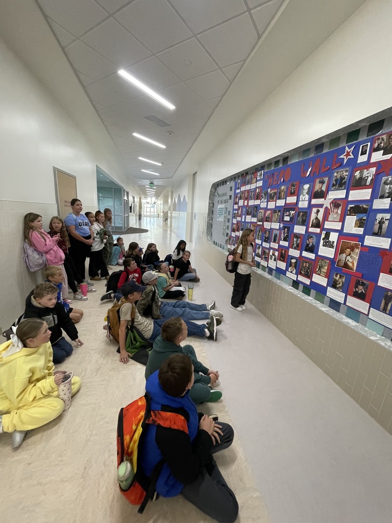 A group of children are sitting on the floor in a brightly lit school hallway, looking up at a large bulletin board titled "HERO WALL" featuring many photos of military service members. One girl stands near the wall, sharing information with the group.