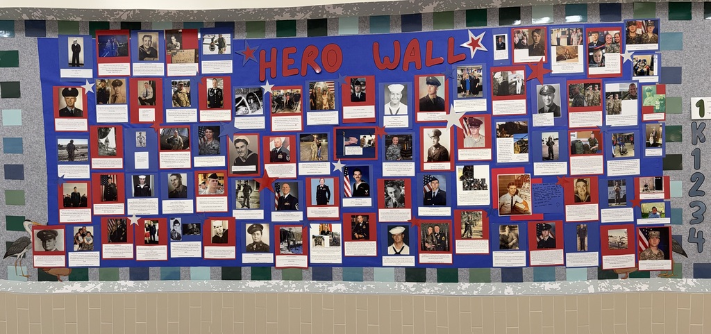 A close-up of a bulletin board in a school hallway titled "HERO WALL" in red and blue letters, covered with dozens of photographs of military service members, many with red and blue borders.
