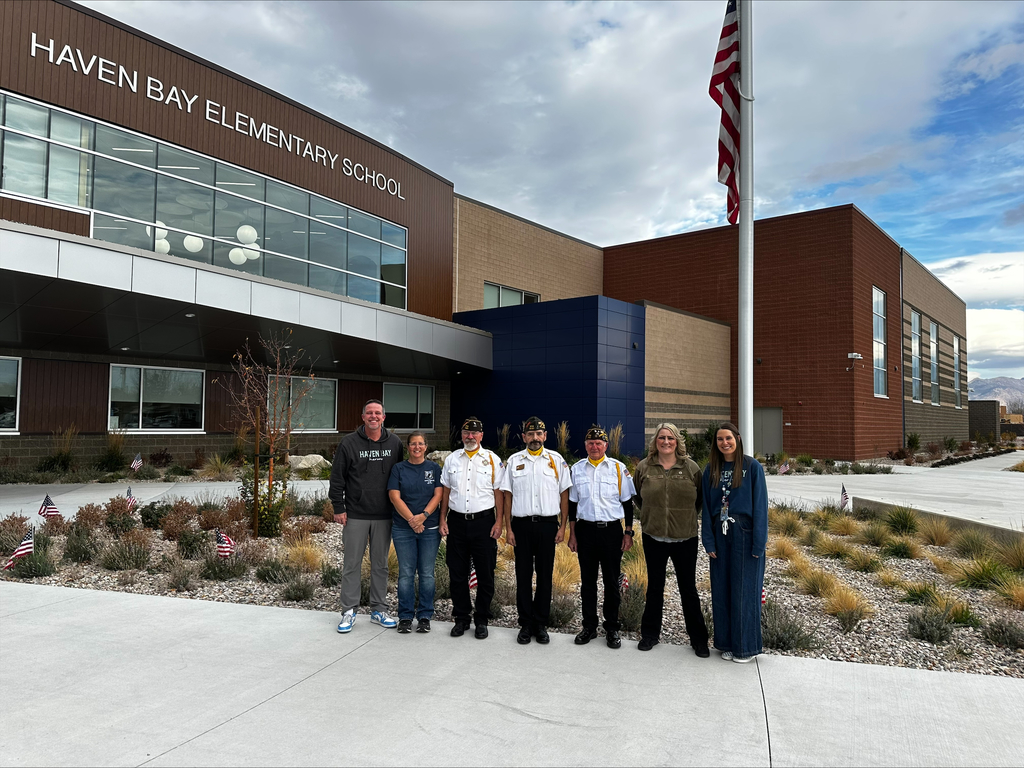 A group of seven adults, including four men in Veterans of Foreign Wars (VFW) uniforms, are standing in front of HAVEN BAY ELEMENTARY SCHOOL next to a flagpole flying the American flag. Small American flags are planted in the flowerbeds.