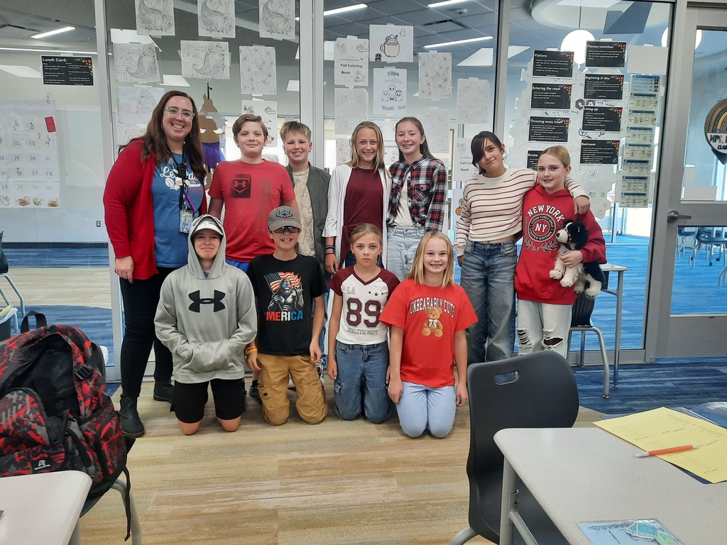 A group of about ten 6th-grade students and a female teacher are posing indoors in a classroom with large windows. The teacher and many of the students are wearing red or patriotic colors. A backpack and a desk with papers are visible in the foreground.