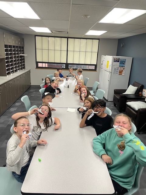 A group of about twelve 4th-grade students is sitting around two long tables in a classroom or small meeting room, all smiling and holding ice cream sandwiches up to their mouths as they eat. They look excited and happy. The room has storage cubbies along the left wall, a refrigerator, and large windows in the background.