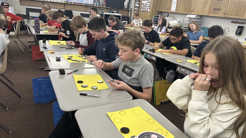 a 6th-grade classroom with students sitting at individual desks. Each student has a stack of Oreo cookies and a bright yellow paper with drawn circles on it. The students are carefully twisting open the Oreos and using tools or their hands to scrape the white cream to represent the different phases of the moon (e.g., crescent, half moon, gibbous). Some students are also seen eating the leftover cookies or cream. The classroom has typical shelving and storage in the background.