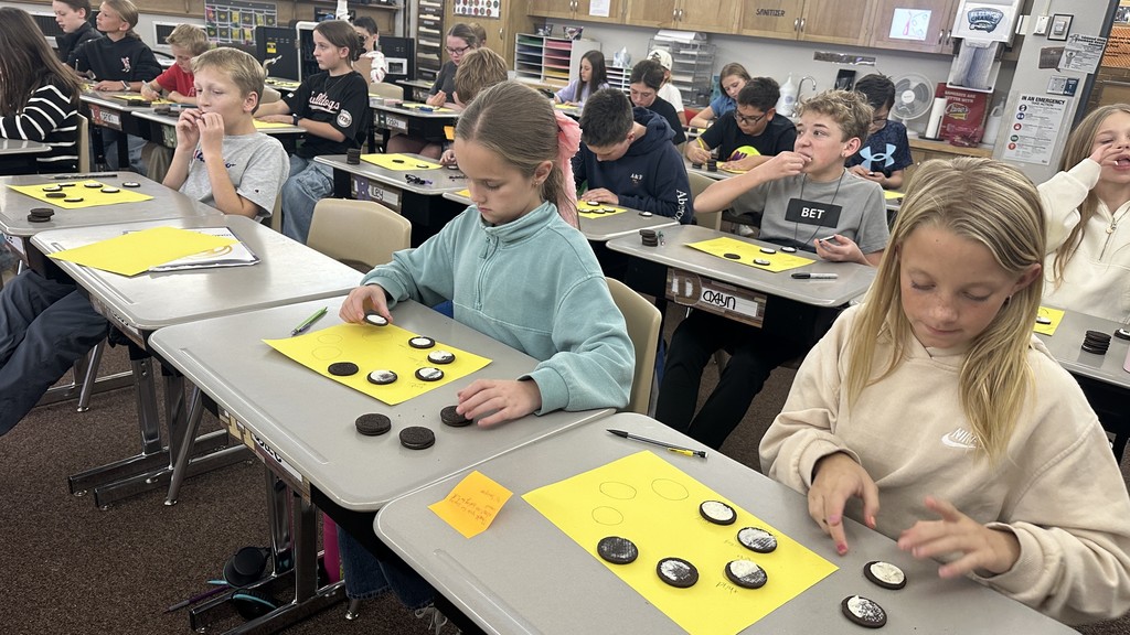 a 6th-grade classroom with students sitting at individual desks. Each student has a stack of Oreo cookies and a bright yellow paper with drawn circles on it. The students are carefully twisting open the Oreos and using tools or their hands to scrape the white cream to represent the different phases of the moon (e.g., crescent, half moon, gibbous). Some students are also seen eating the leftover cookies or cream. The classroom has typical shelving and storage in the background.