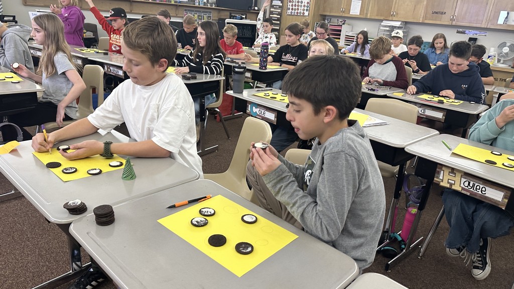 a 6th-grade classroom with students sitting at individual desks. Each student has a stack of Oreo cookies and a bright yellow paper with drawn circles on it. The students are carefully twisting open the Oreos and using tools or their hands to scrape the white cream to represent the different phases of the moon (e.g., crescent, half moon, gibbous). Some students are also seen eating the leftover cookies or cream. The classroom has typical shelving and storage in the background.