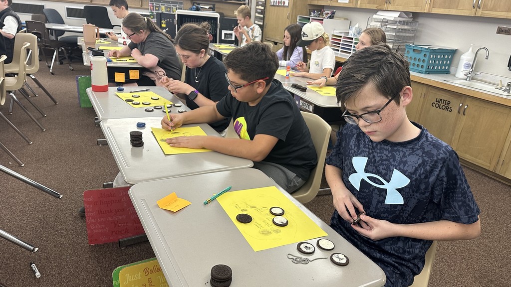 a 6th-grade classroom with students sitting at individual desks. Each student has a stack of Oreo cookies and a bright yellow paper with drawn circles on it. The students are carefully twisting open the Oreos and using tools or their hands to scrape the white cream to represent the different phases of the moon (e.g., crescent, half moon, gibbous). Some students are also seen eating the leftover cookies or cream. The classroom has typical shelving and storage in the background.