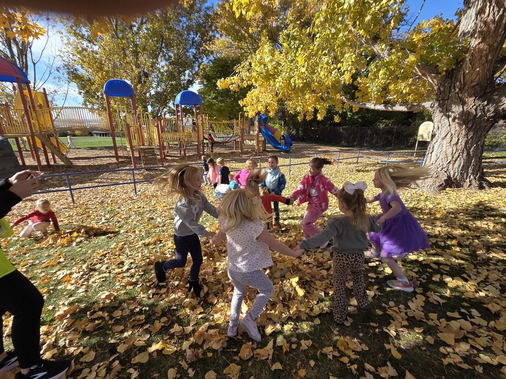 A group of elementary school-aged children holding hands and playing a game, possibly "Ring Around the Rosie," outdoors on a sunny autumn day. They are encircled by yellow leaves covering the grass under a large tree with bright golden foliage. A playground with slides and climbing structures is visible in the background.