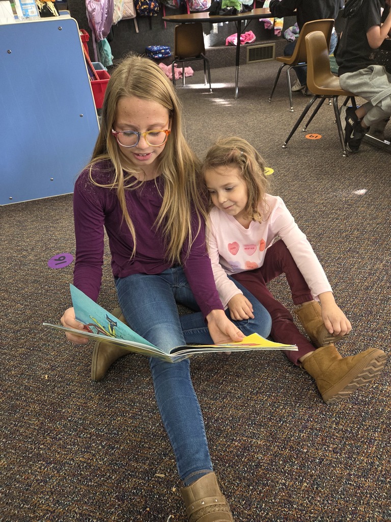 A photograph showing two female students sitting on a carpeted classroom floor. An older student with long blonde hair and glasses is holding an open book and reading to a younger student who is sitting close beside her and looking intently at the pages.