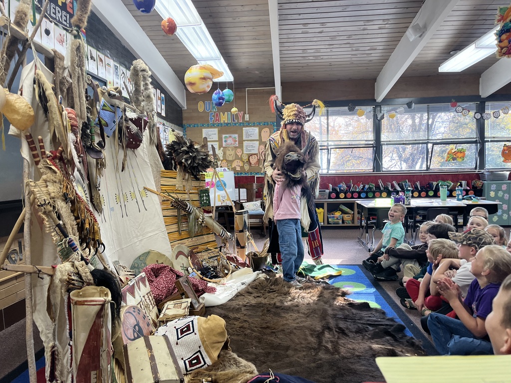 A series of photographs showing a cultural presenter, dressed in traditional regalia, teaching a group of elementary students sitting on a classroom floor. The presenter is surrounded by artifacts and hides, including a large display resembling a tepee on the left. Several images show the presenter interacting directly with individual students or students raising their hands to ask questions.
