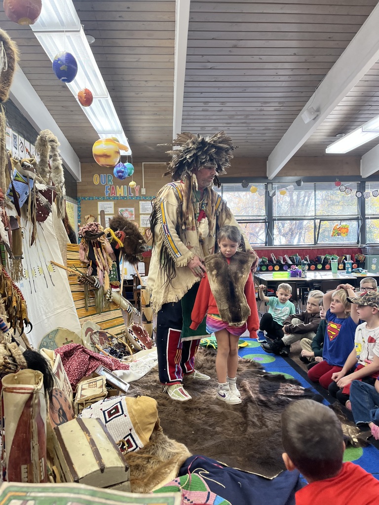 A series of photographs showing a cultural presenter, dressed in traditional regalia, teaching a group of elementary students sitting on a classroom floor. The presenter is surrounded by artifacts and hides, including a large display resembling a tepee on the left. Several images show the presenter interacting directly with individual students or students raising their hands to ask questions.