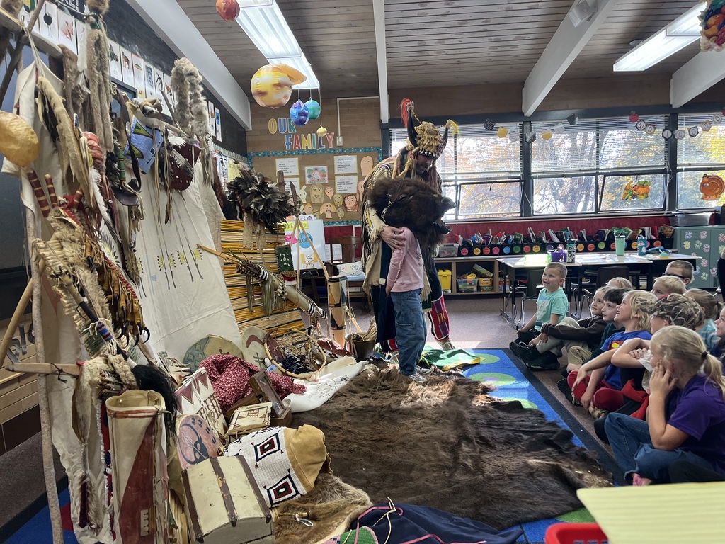 A series of photographs showing a cultural presenter, dressed in traditional regalia, teaching a group of elementary students sitting on a classroom floor. The presenter is surrounded by artifacts and hides, including a large display resembling a tepee on the left. Several images show the presenter interacting directly with individual students or students raising their hands to ask questions.