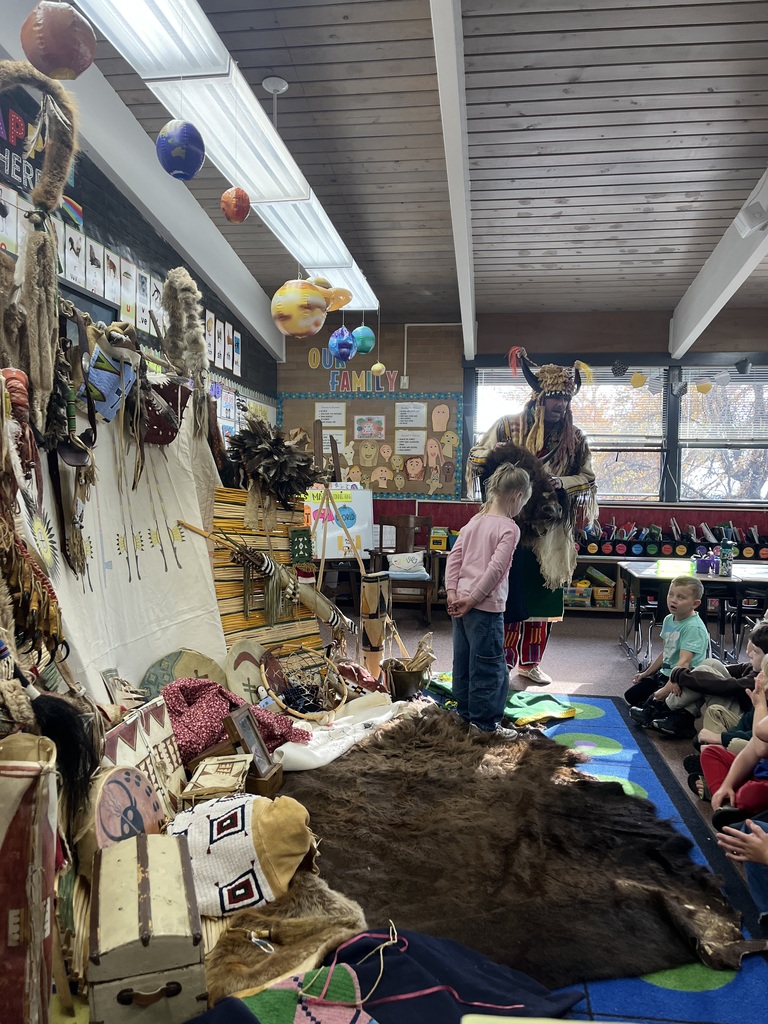 A series of photographs showing a cultural presenter, dressed in traditional regalia, teaching a group of elementary students sitting on a classroom floor. The presenter is surrounded by artifacts and hides, including a large display resembling a tepee on the left. Several images show the presenter interacting directly with individual students or students raising their hands to ask questions.