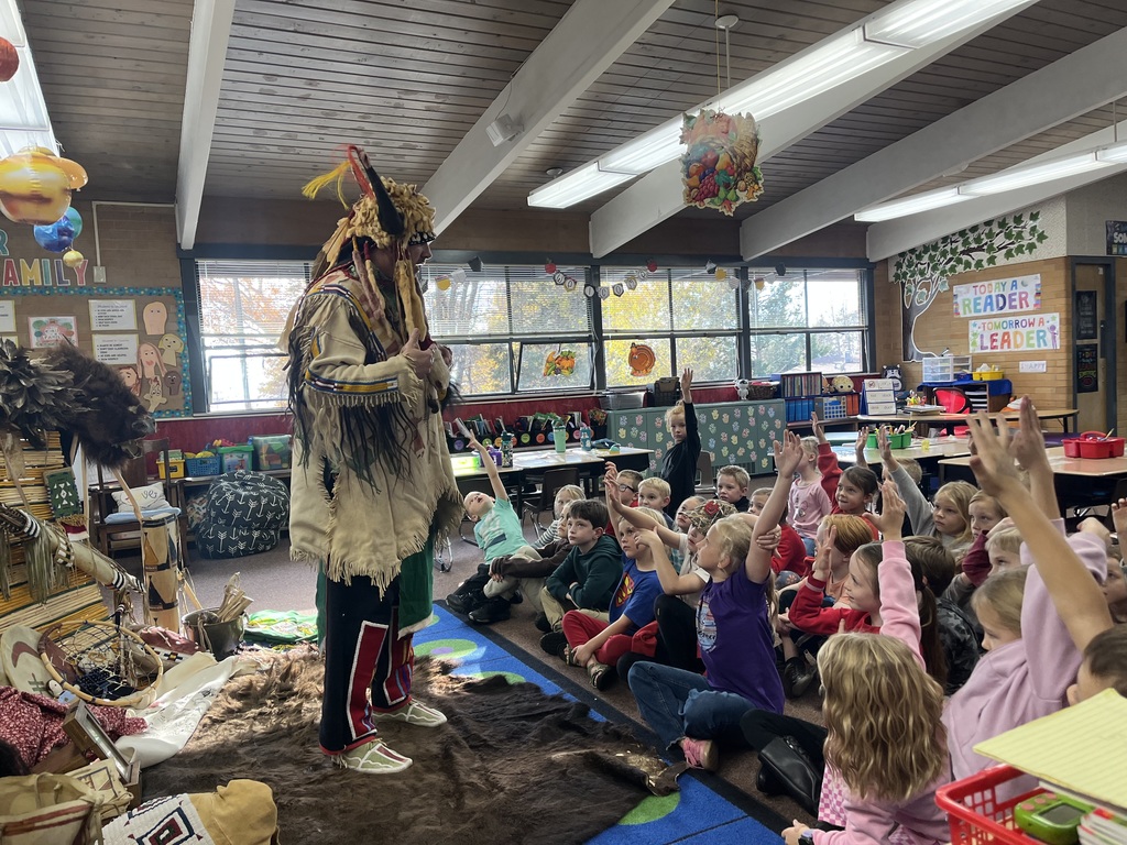 A series of photographs showing a cultural presenter, dressed in traditional regalia, teaching a group of elementary students sitting on a classroom floor. The presenter is surrounded by artifacts and hides, including a large display resembling a tepee on the left. Several images show the presenter interacting directly with individual students or students raising their hands to ask questions.