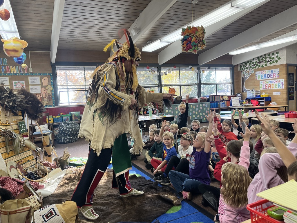 A series of photographs showing a cultural presenter, dressed in traditional regalia, teaching a group of elementary students sitting on a classroom floor. The presenter is surrounded by artifacts and hides, including a large display resembling a tepee on the left. Several images show the presenter interacting directly with individual students or students raising their hands to ask questions.