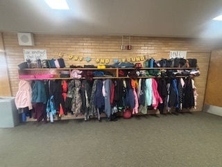 A long lost and found shelf and coat rack in a school hallway, overflowing with numerous children's jackets, coats, sweatshirts, lunchboxes, and water bottles. The items are neatly hung on hangers and piled on the shelf, with a 'LOST AND FOUND' banner in colorful letters above