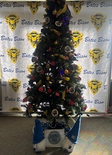 A picture of Bates Elementary's Giving Tree.  A Christmas tree  decorated in Bison ornaments, blue and yellow, and red Christmas tags.  There are two gnomes in front of the tree with blue hats and gloves next to a sign that says Giving Tree.  the back ground is of a white canvas photo backdrop with yellow and blue bison and Bates Bison.