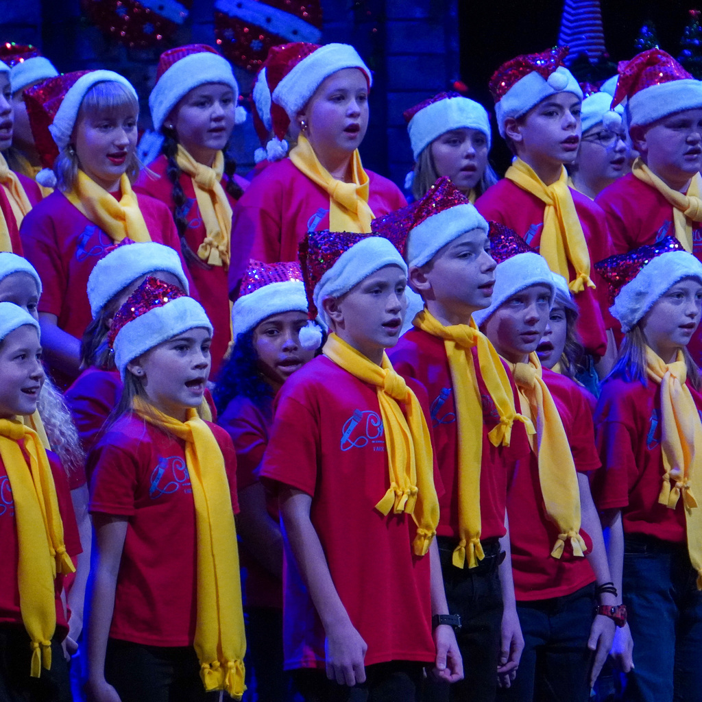 Multiple children in matching shirts , yellow scarfs, and santa hats sing on a stage