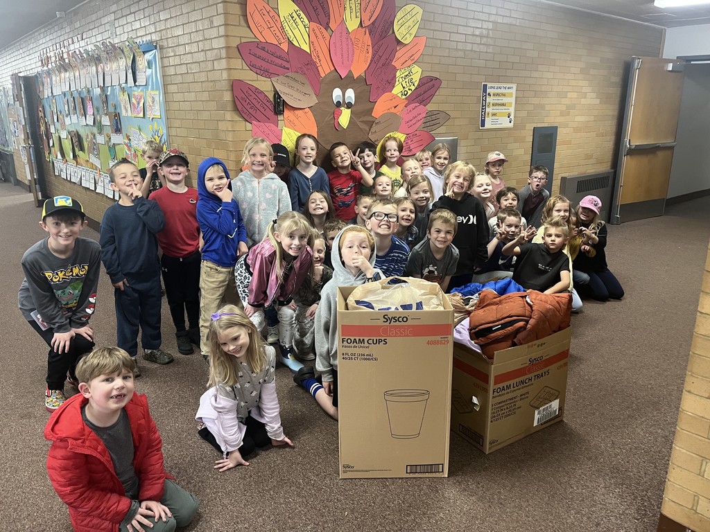 A large group of approximately 25 first-grade students posing in a school hallway with several boxes full of donated coats and blankets. Behind them is a large bulletin board decoration of a turkey whose feathers are made of colorful cutouts with writing on them.