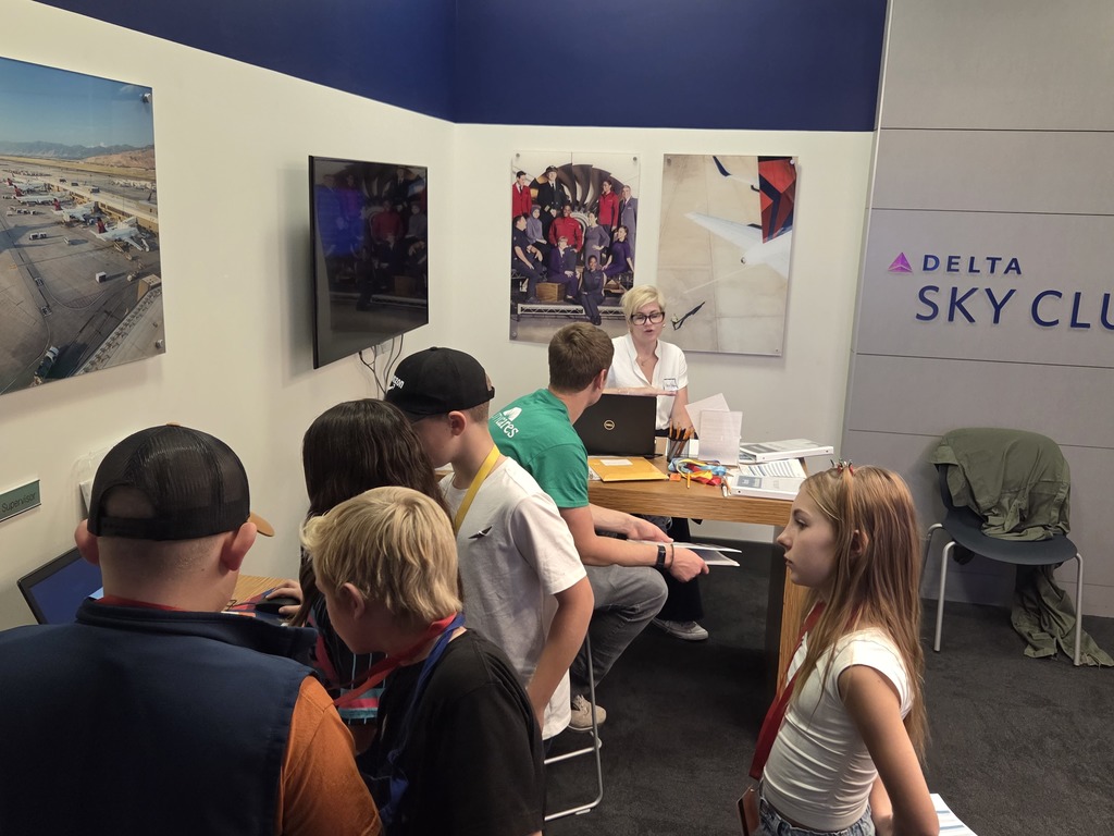 A group of students gather around a desk in a simulated airport or airline office, labeled "DELTA SKY CLUB". An adult with blonde hair and glasses is seated at the desk working on a laptop and talking to the children. Airline-themed artwork and photography decorate the walls.