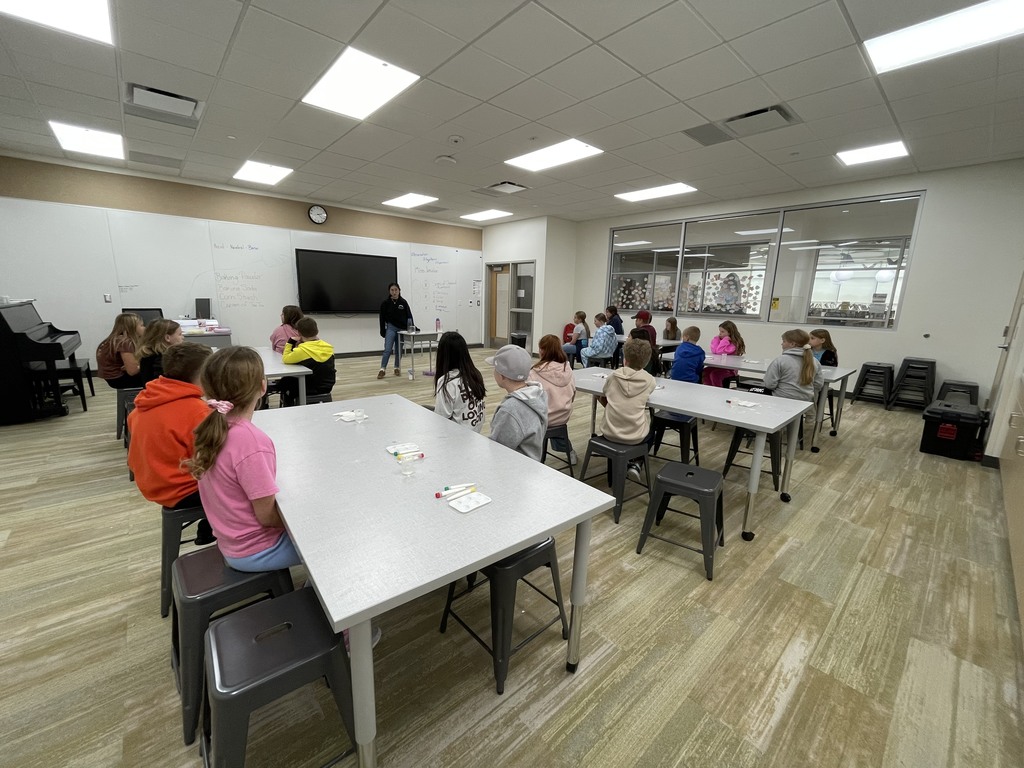 A wide view of a modern classroom with about 15 students seated at rectangular white tables, looking toward the front where a teacher is standing near a large monitor and whiteboard. The students are participating in a hands-on science lesson with materials spread out on the tables.