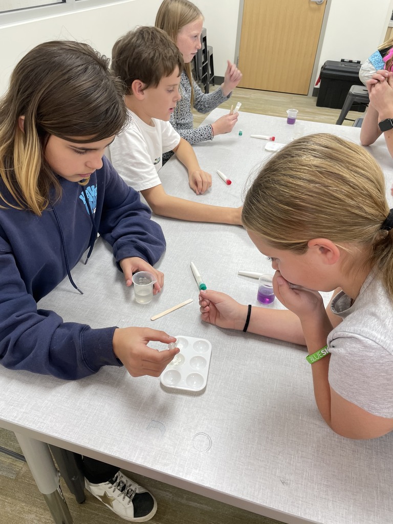 Close-up of three students, two girls and one boy, conducting a science experiment at a white table. The girl on the left is holding a small vial and using a wooden stick with a white multi-well plate. The girl on the right is looking closely at a small vial containing a purple liquid. Test tubes, pipettes, and small cups are on the table.