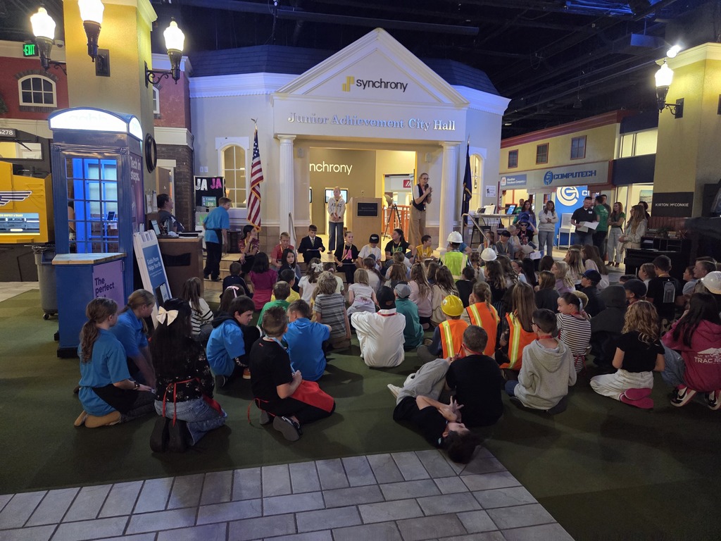 A large group of children and adults are gathered on the floor in front of a miniature building facade labeled "synchrony Junior Achievement City Hall". A few adults stand on a stage near the entrance, addressing the audience. The setting is an indoor educational center designed to look like a small city.