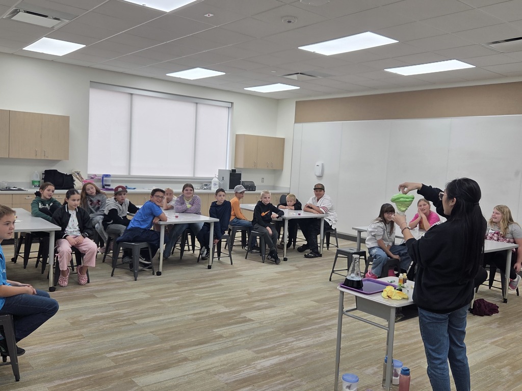 A diverse group of approximately 15 middle school students sitting at white tables, watching a demonstration by a teacher. The teacher is standing on the right, holding a large beaker containing a bright, neon green liquid, demonstrating a chemical reaction to the class. The classroom has beige cabinets, a white board, and patterned carpet.