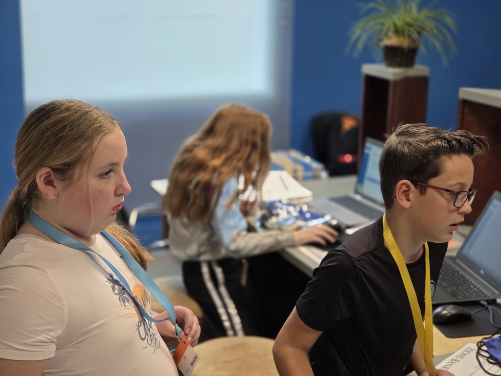 Two students, a girl with blonde hair in a ponytail and a boy in glasses, look intently at computer screens in a simulated office or classroom setting. A third student with long red hair is blurred in the background working on a laptop.